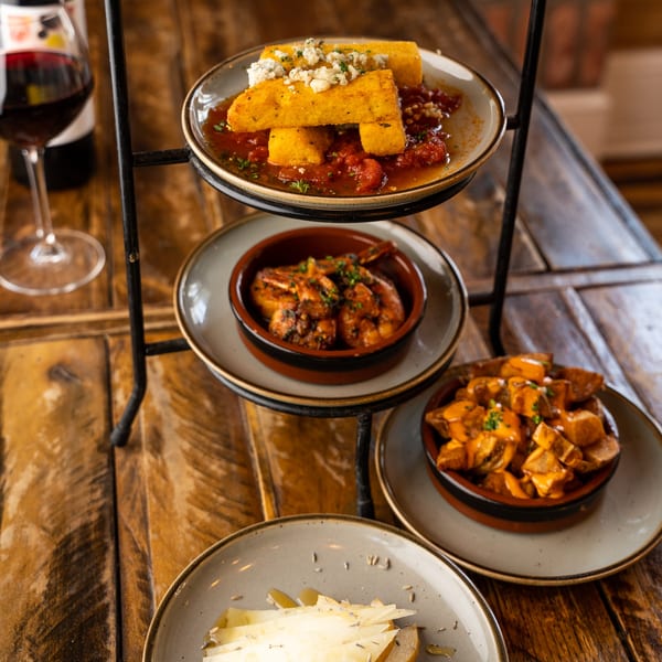A tiered stand with various New American tapas, including polenta, shrimp, potatoes, and cheese, with red wine.