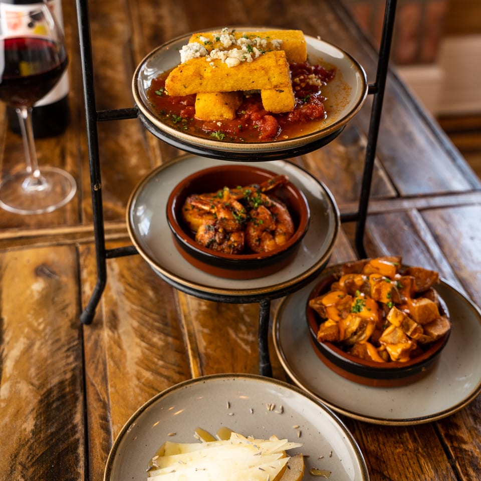 A tiered stand with various New American tapas, including polenta, shrimp, potatoes, and cheese, with red wine.
