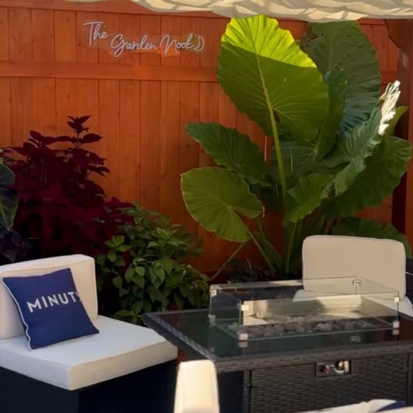 Outdoor restaurant lounge with white seating, a fire pit, tropical plants, and a wooden wall sign reading “The Garden Marks.”