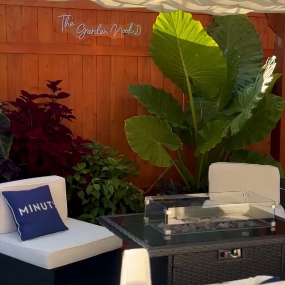 Outdoor restaurant lounge with white seating, a fire pit, tropical plants, and a wooden wall sign reading “The Garden Marks.”