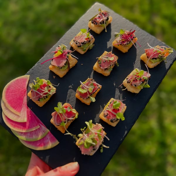 A hand holds a slate tray of tuna tartare appetizers with watermelon radish slices, against a green background.