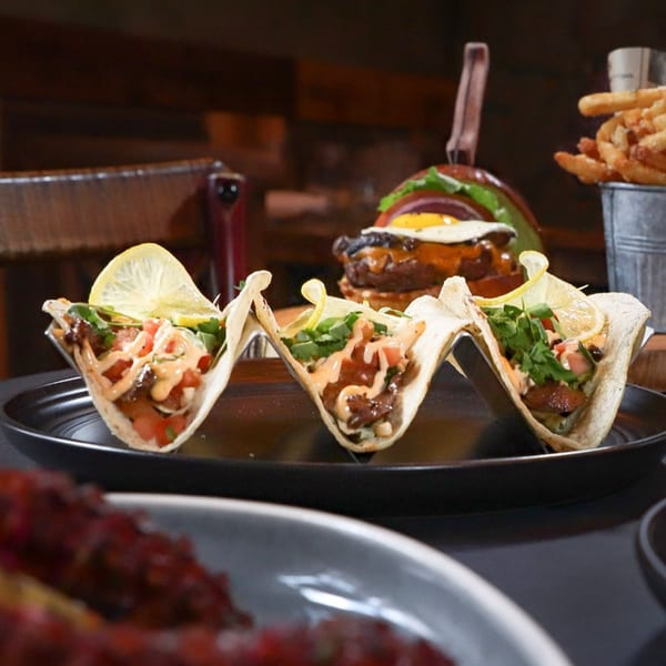 Three tacos with meat, salsa, and lemon wedges, with a burger and fries in the background.