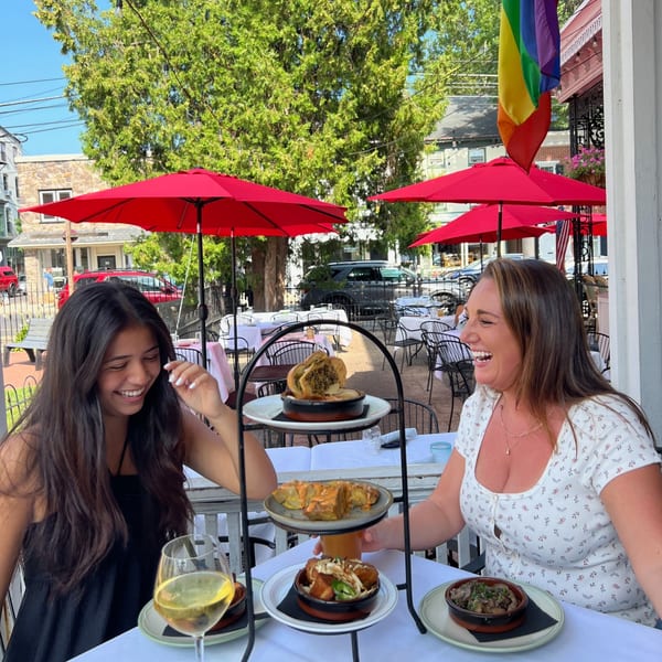 Two women laughing at an outdoor table with a tiered stand of New American dishes and drinks.