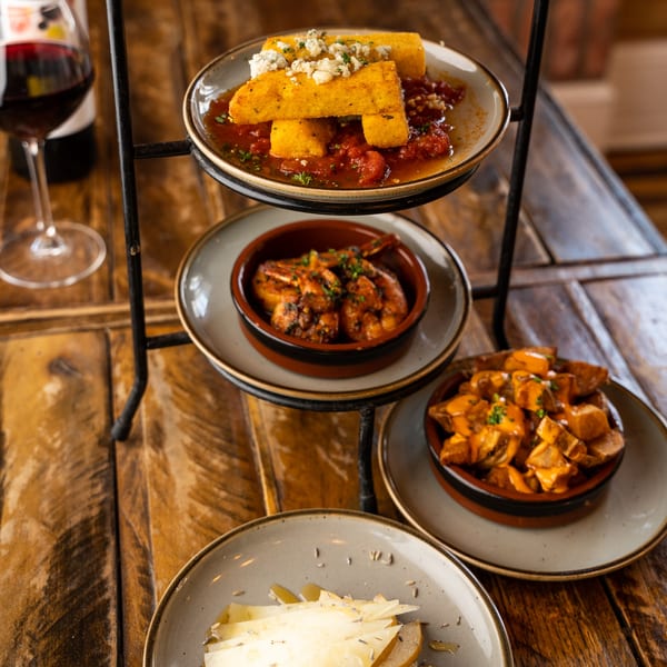 A tiered stand with polenta, shrimp, potatoes, and cheese with pears, alongside a glass of red wine.