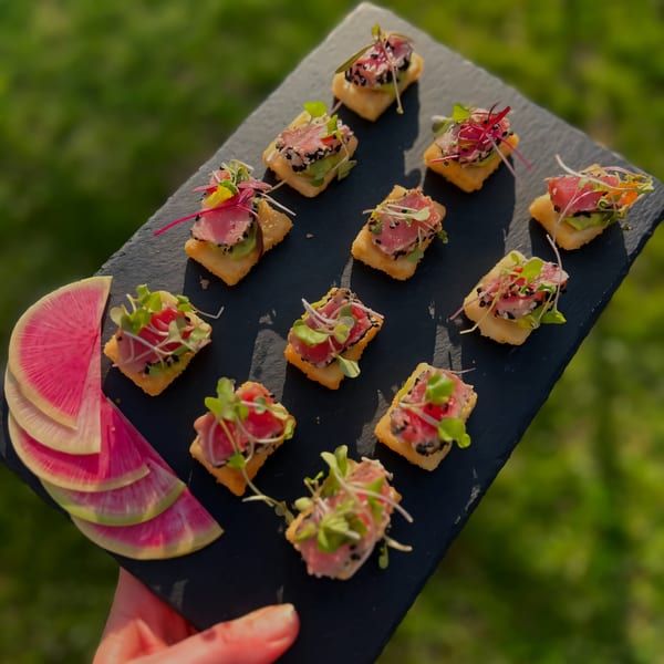 A hand holds a black slate tray of tuna appetizers with watermelon radish slices, against a green background.