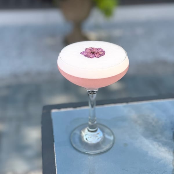 Pink cocktail in a stemmed coupe glass, topped with a delicate purple flower, sits on a blue tabletop.