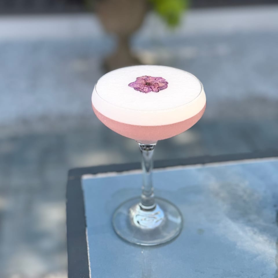 Pink cocktail in a stemmed coupe glass, topped with a delicate purple flower, sits on a blue tabletop.