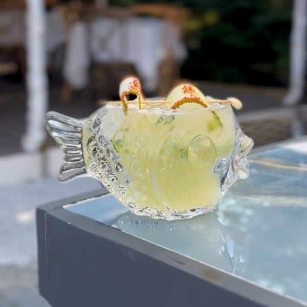 Pale yellow cocktail garnished with citrus slices and herbs, served in an ornate fish-shaped glass on a reflective table.