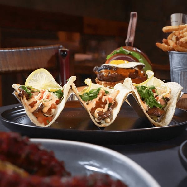 Three tacos with lemon wedges, a burger, and fries in a bucket on a dark table.