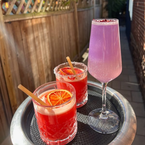 Three vibrant cocktails, two red with orange slices and one shimmering purple, on a tray outdoors.