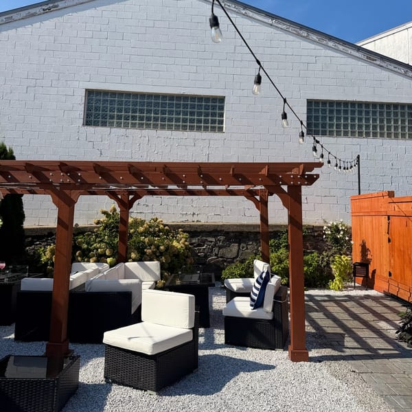 Outdoor restaurant patio with cushioned wicker seating beneath a wooden pergola, string lights, greenery, and a white brick wall.