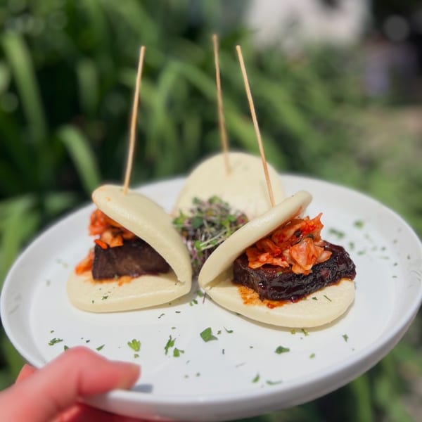 Three bao buns with dark meat and kimchi, garnished with microgreens on a white plate.
