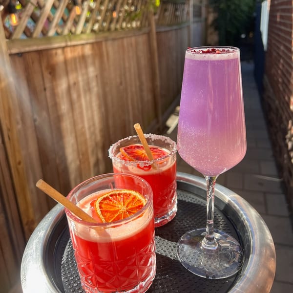 Three vibrant cocktails, two red with orange slices and one sparkling purple, on a tray outdoors.