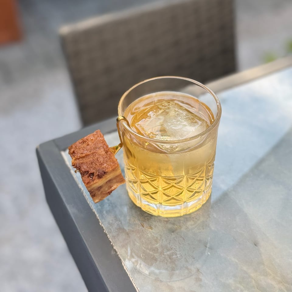 Golden cocktail with ice in a patterned glass, served alongside a rectangular chocolate dessert on a dark tray.