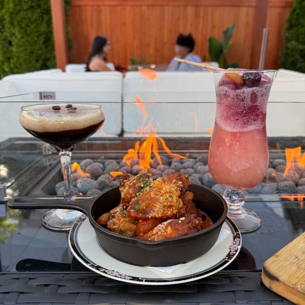 Skillet of saucy fried chicken served beside dark and pink cocktails at an outdoor firepit table.